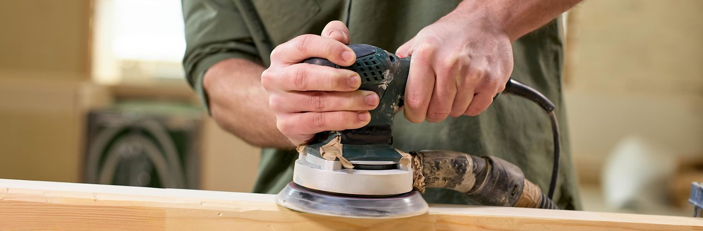 Ein Mann arbeitet mit einer Schleifmaschine. Der Bildausschnitt zeigt wie ein Mann mit einer runden Schleifmaschine einen Holzbalken schleift.