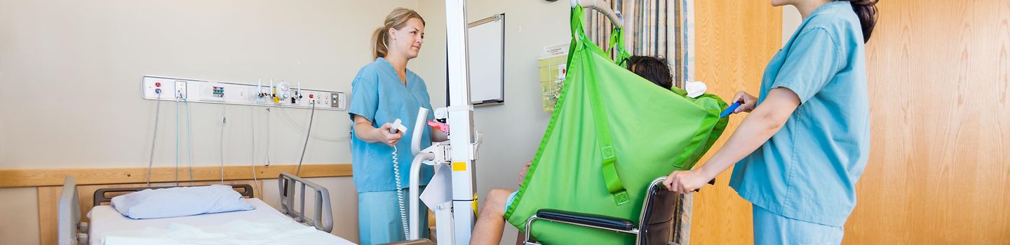 In einem Krankenhauszimmer benutzen zwei Krankenschwestern eine Hebehilfe, um den Patienten in einen Rollstuhl zu setzen In einem Krankenhauszimmer benutzen zwei Krankenschwestern eine Hebehilfe, um den Patienten in einen Rollstuhl zu setzen