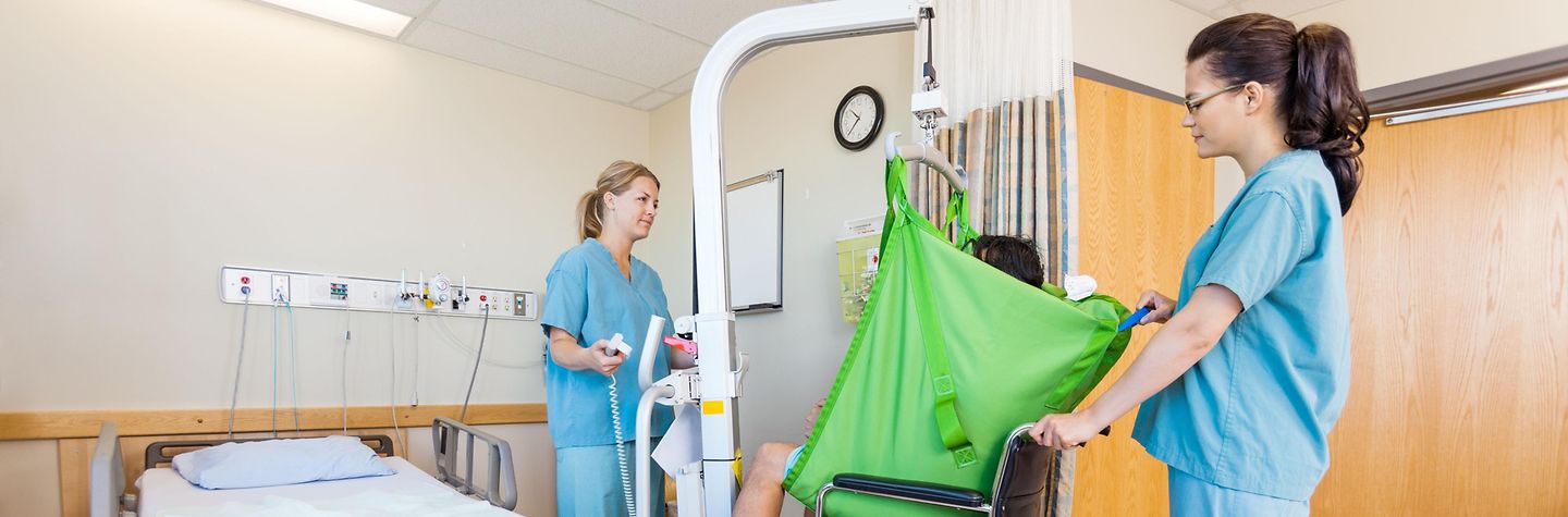 In einem Krankenhauszimmer benutzen zwei Krankenschwestern eine Hebehilfe, um den Patienten in einen Rollstuhl zu setzen