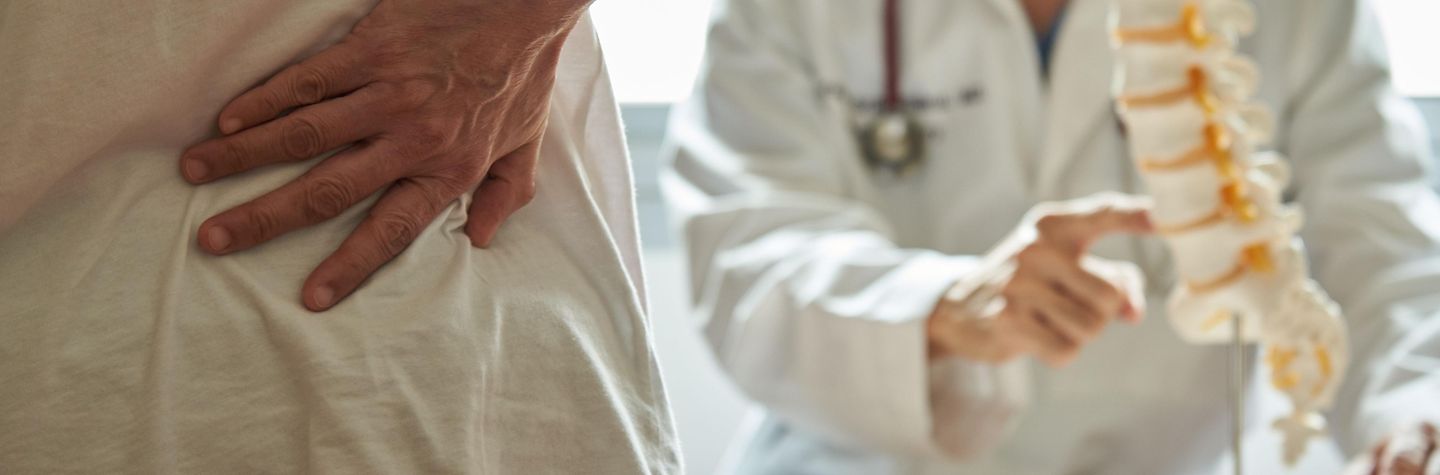 Ein Arzt erklärt einem Patienten anhand eines Modells, woher der Schmerz kommt. Ein Arzt erklärt an einem Modell einem Patienten, der sich den Rücken hält, woher der Schmerz kommt