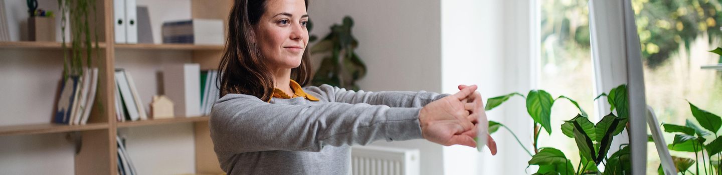 Junge Frau arbeitet zu Hause an einem Notebook. Sie streckt die Arme nach vorne und macht Dehnungsübungen.