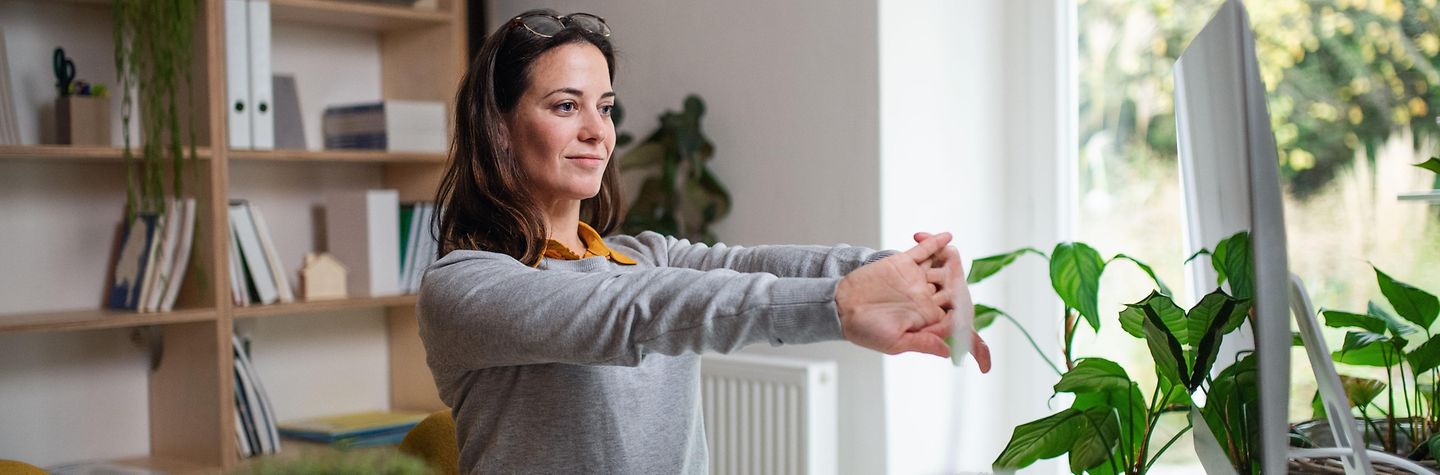 Homeoffice Junge Frau arbeitet zu Hause an einem Notebook. Sie streckt die Arme nach vorne und macht Dehnungsübungen.